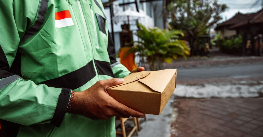 A person in a green jacket delivering a package outdoors. Urban setting, daytime.